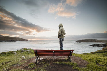 Spain, Ferrol, man wearing hooded jacket standing on a bench at the coast looking at distance - RAEF000904