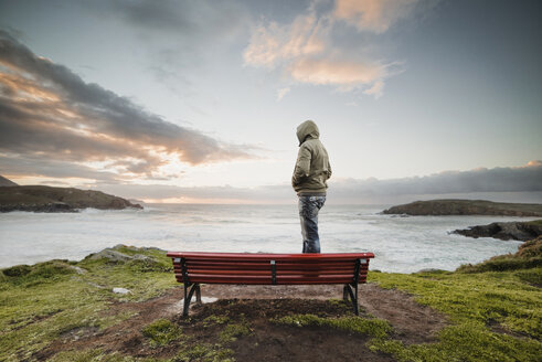 Spain, Ferrol, man wearing hooded jacket standing on a bench at the coast looking at distance - RAEF000904