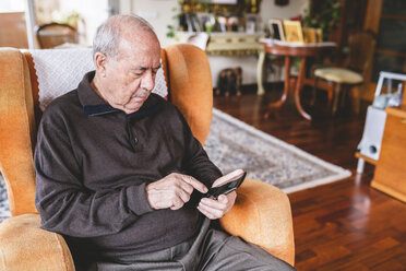 Älterer Mann sitzt zu Hause auf einem Sessel und benutzt ein Smartphone - GEMF000744