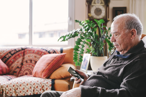 Älterer Mann sitzt im Wohnzimmer und benutzt sein Smartphone, lizenzfreies Stockfoto