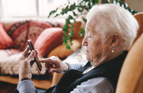 Ältere Frau sitzt im Wohnzimmer und benutzt ihr Smartphone, lizenzfreies Stockfoto