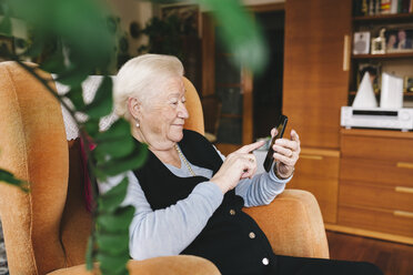 Senior woman sitting in the living room using smartphone - GEMF000754