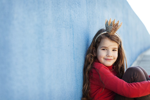 Portrait of smiling ittle girl with a crown leaning against blue wall stock photo