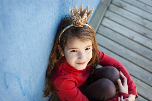 Porträt eines kleinen Mädchens mit einer Krone, das an einer blauen Wand lehnt und zur Kamera aufschaut - VABF000209