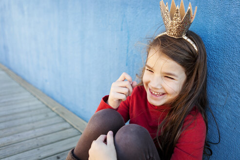 Portrait of laughing little girl with a crown leaning against blue wall - VABF000210