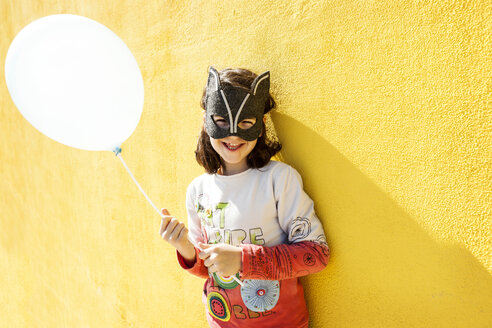 Portrait of laughing little girl with balloon wearing animal mask in front of yellow wall - VABF000214