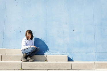 Portrait of smiling little girl sitting on steps in front of blue wall - VABF000216