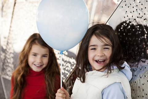 Porträt eines lachenden kleinen Mädchens mit Ballon und Schwester im Hintergrund, lizenzfreies Stockfoto