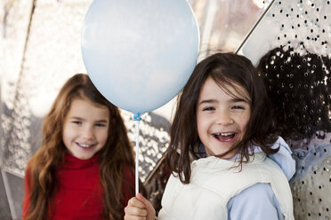 Porträt eines lachenden kleinen Mädchens mit Ballon und Schwester im Hintergrund - VABF000218