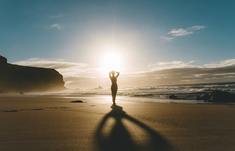 Frau stehend am Strand mit Händen auf dem Kopf bei Sonnenuntergang, lizenzfreies Stockfoto