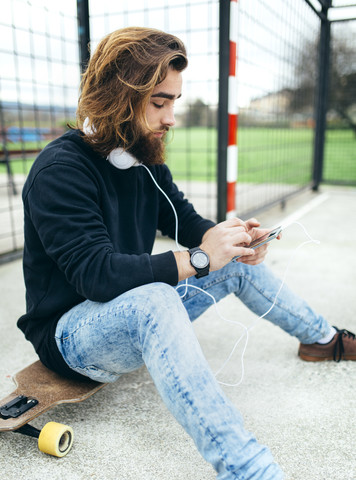 Bärtiger junger Skateboarder mit Smartphone und Kopfhörern, lizenzfreies Stockfoto