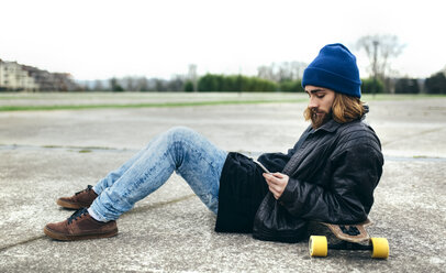 Junger Skateboarder sitzt auf dem Boden und schaut auf sein Smartphone - MGOF001465