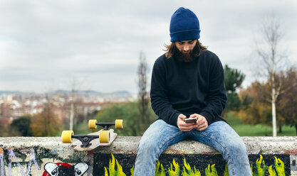 Bärtiger junger Skateboarder sitzt an einer Wand und schaut auf sein Smartphone - MGOF001470