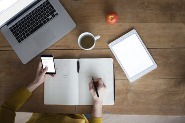 Woman at wooden table using smartphone and writing in notebook - RBF004101