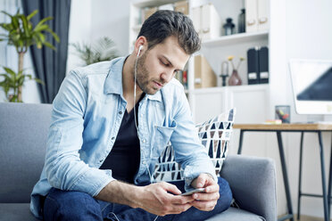 Junger Mann zu Hause mit Ohrstöpseln und Blick auf sein Smartphone - SEGF000438