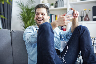 Happy young man at home with smartphone and earbuds - SEGF000441