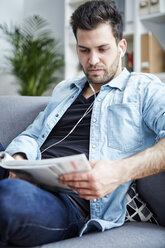 Junger Mann zu Hause mit Kopfhörern beim Lesen einer Zeitschrift - SEGF000443