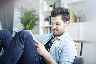 Junger Mann zu Hause mit Kopfhörern beim Lesen einer Zeitschrift - SEGF000444