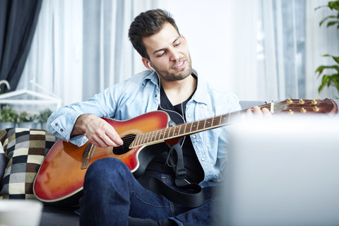 Junger Mann sitzt zu Hause auf der Couch und spielt Gitarre, lizenzfreies Stockfoto