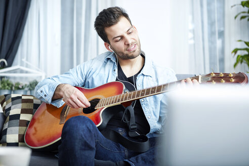 Young man at home sitting on couch playing guitar - SEGF000447