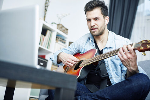Young man at home sitting on couch playing guitar and looking at laptop - SEGF000451