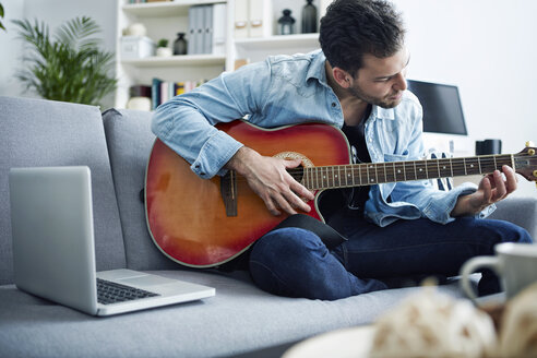 Young man at home sitting on couch playing guitar next to laptop - SEGF000452
