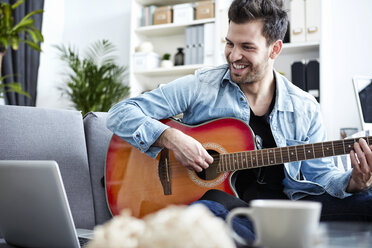 Junger Mann sitzt zu Hause auf der Couch, spielt Gitarre und schaut auf den Laptop - SEGF000453