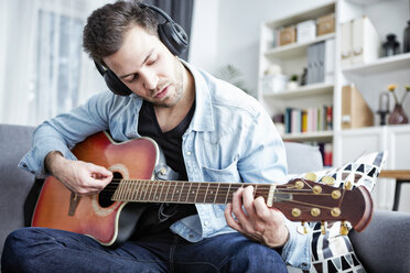 Junger Mann sitzt zu Hause auf der Couch, trägt Kopfhörer und spielt Gitarre - SEGF000454