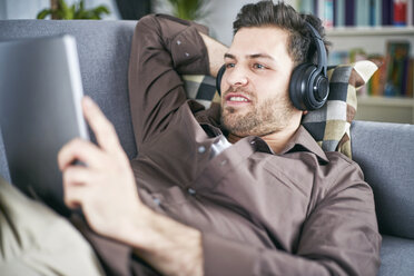 Young man lying on couch with headphones looking at digital tablet - SEGF000482