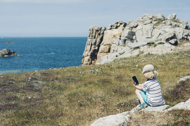 Frankreich, Bretagne, Cote de Granit Rose, Junge sitzt mit Handy an der Küste - MJF001764