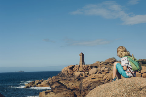 Frankreich, Bretagne, Cote de Granit Rose, Junge sitzt auf Felsen an der Küste, lizenzfreies Stockfoto