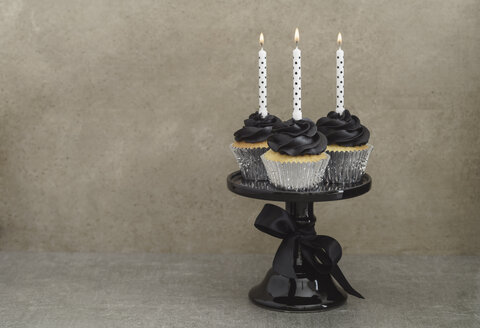 Drei Tassenkuchen mit schwarzer Buttercreme und brennenden Kerzen auf einem Tortenständer - ECF001859
