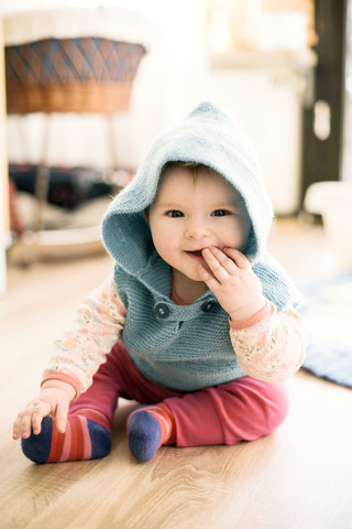 Portrait of smiling baby girl wearing hooded jacket sitting on the floor with finger in mouth stock photo