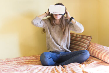 Junge Frau mit Virtual-Reality-Brille, die zu Hause auf ihrem Bett sitzt - SKCF000075