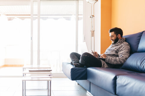 Bärtiger junger Mann arbeitet zu Hause entspannt auf der Couch sitzend, mit Laptop und Handy - JRFF000471