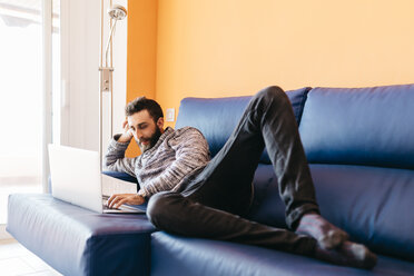 Bärtiger junger Mann arbeitet zu Hause entspannt auf der Couch liegend, mit Laptop - JRFF000473