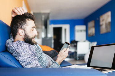 Bärtiger junger Mann arbeitet zu Hause entspannt auf der Couch sitzend, mit Laptop und Handy - JRFF000474