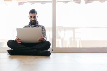 Bärtiger junger Mann arbeitet entspannt zu Hause, sitzt auf dem Boden und benutzt einen Laptop - JRFF000476