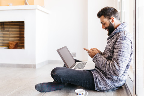 Bärtiger junger Mann, der entspannt zu Hause arbeitet, auf dem Boden sitzend, mit Laptop und Handy, lizenzfreies Stockfoto