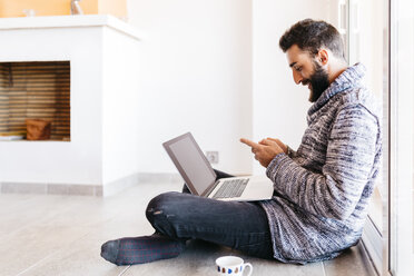 Bearded young man working at home relaxed, sitting on the floor, using laptop and mobile - JRFF000477