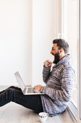 Bearded young man working at home relaxed, sitting on the floor, using laptop - JRFF000478