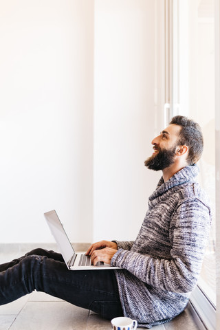 Bärtiger junger Mann arbeitet entspannt zu Hause, sitzt auf dem Boden und benutzt einen Laptop, lizenzfreies Stockfoto
