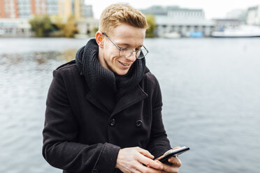 Irland, Dublin, lächelnder junger Mann, der vor dem Wasser auf sein Smartphone schaut - BOYF000103