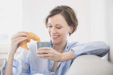 Porträt einer entspannten Frau auf der Couch, die ein Croissant isst und Kaffee trinkt - FMKF002412