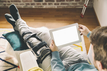 Injured man with knee brace using a tablet computer at home - MFF002726