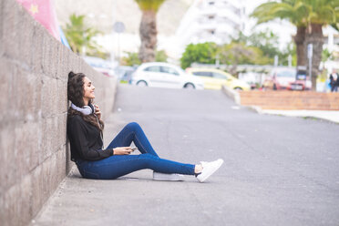 Spain, Tenerife, smiling teenage girl with headphones and smartphone sitting on the ground - SIPF000217