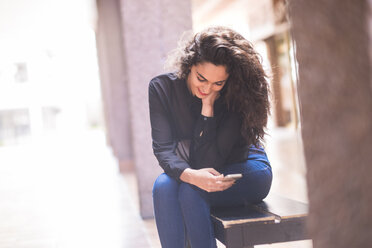 Lächelndes Teenager-Mädchen sitzt auf einer Bank und schaut auf ihr Smartphone - SIPF000224