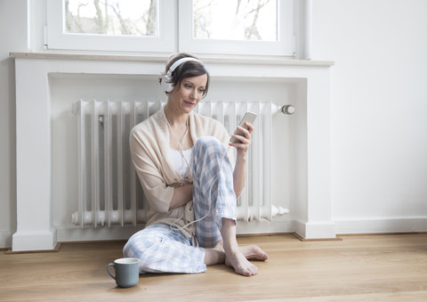 Frau zu Hause auf dem Boden sitzend mit Handy und Kopfhörern, lizenzfreies Stockfoto