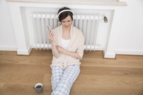 Frau zu Hause auf dem Boden sitzend mit Handy und Kopfhörern, lizenzfreies Stockfoto