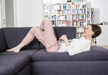 Happy woman lying on couch using laptop - FMKF002428